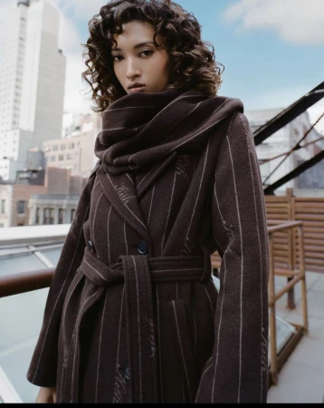 Model with dark curly hair wearing a brown belted coat on a rooftop overlooking city buildings against a blue sky.