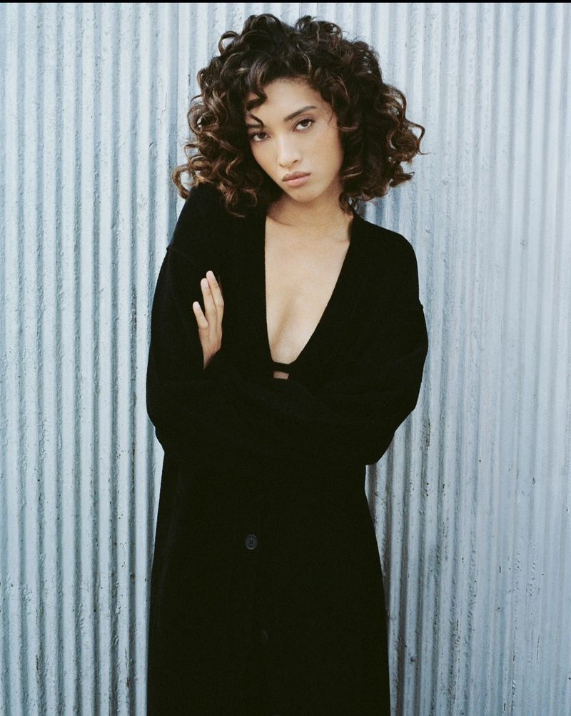 Model with dark curly hair wearing a long black coat against a corrugated metal wall