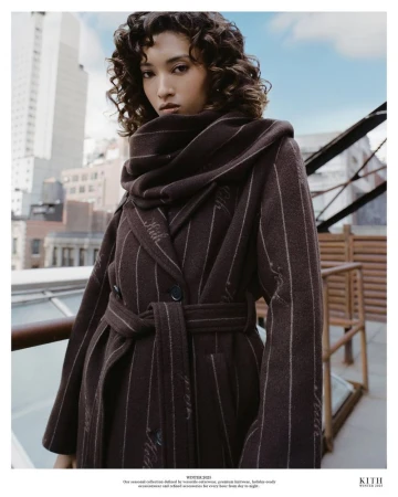 Model Aqua Parios wearing a belted dark brown pinstripe coat and matching scarf on an urban rooftop terrace.