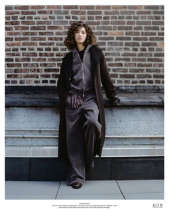 Aqua Parios wearing a long dark overcoat, zip hoodie, and wide-leg trousers against a brick wall for Kith Women Winter 2025.