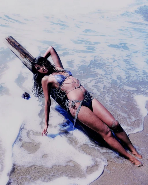 A woman in a bathing suit reclines against driftwood on a sandy beach with waves lapping at the shore in the sunlight.