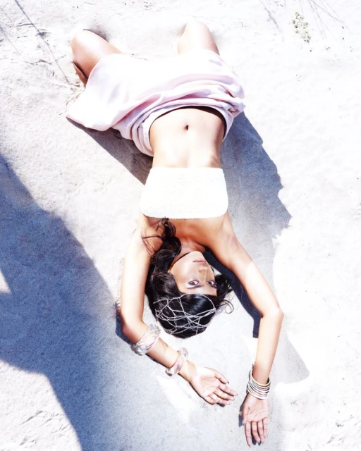Brunette woman in white tube top and pink draped fabric poses on tan sand. Arms are extended above her head, a headpiece adorns her hair.