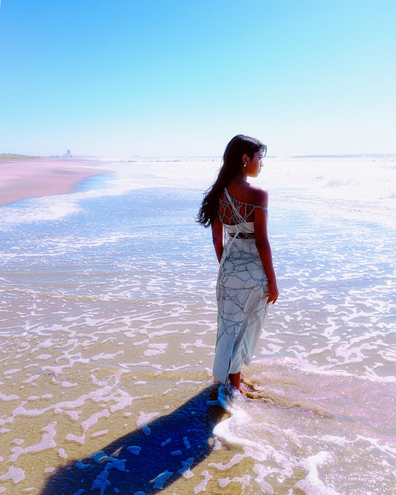 Woman in a light grey dress standing in ocean waves on a sandy beach under a clear sky.