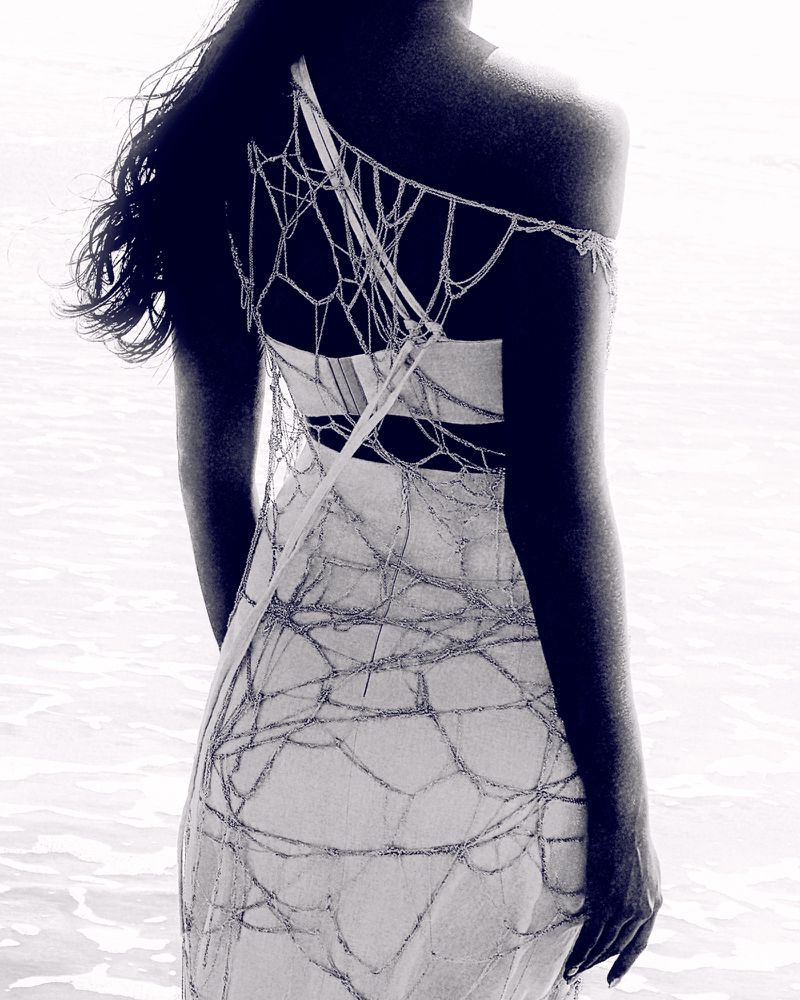 Back view of a slender woman in a light-colored dress with a beaded web design standing in shallow water with a blurred background.