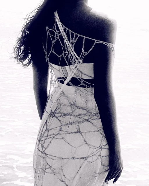 Back view of a slender woman in a light-colored dress with a beaded web design standing in shallow water with a blurred background.