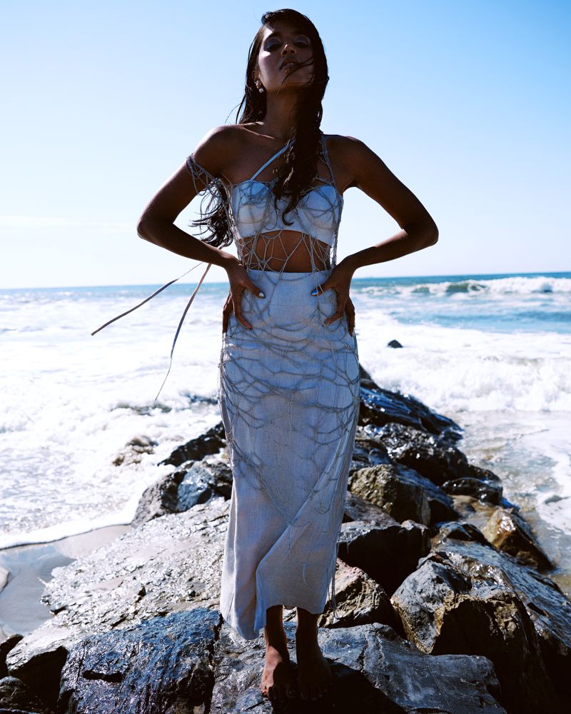 Model standing on rocky shore wearing light blue netted two-piece outfit with ocean waves and clear sky in background.