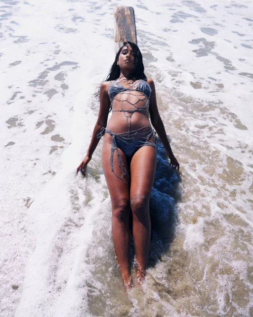 A woman in a swimsuit lies on a log in the ocean surf, with sunlight creating a bright, summery beach vibe. The shot is eye-level.