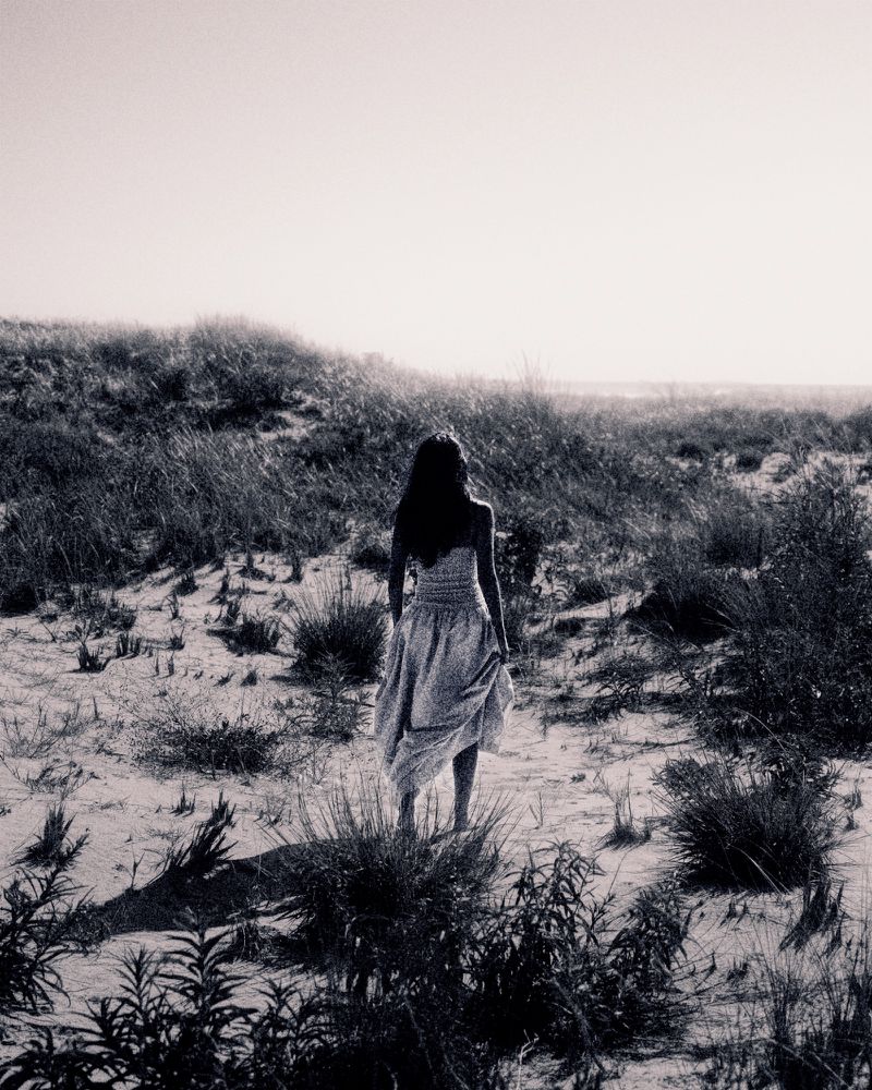 Black and white photograph of a female figure in a light dress walking across a grassy, sandy landscape toward a bright horizon.