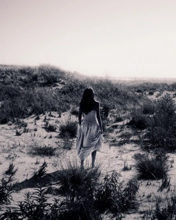 Black and white photograph of a female figure in a light dress walking across a grassy, sandy landscape toward a bright horizon.