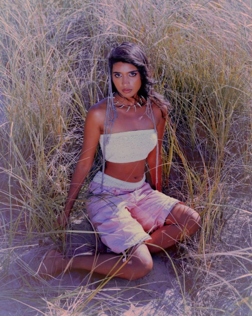 Woman in nature setting, wearing a tube top and shorts, kneeling in the sand surrounded by tall grasses, adorned with necklaces.