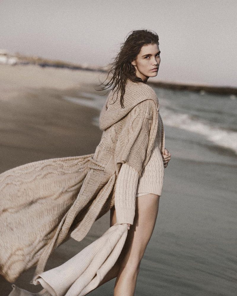 Woman in knitwear poses on a beach with light tan sand and ocean waves. The subject is wearing a long cardigan and has windswept hair.