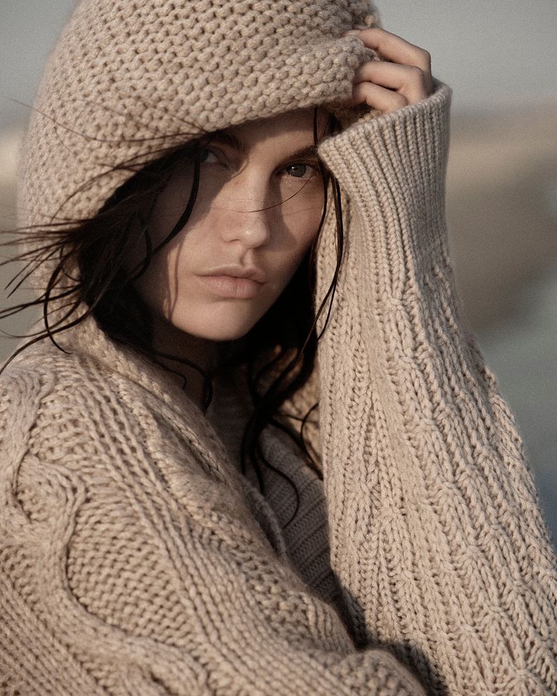 Close-up of an adult female wearing a tan hooded knit sweater with a neutral outdoor background, portrait orientation, eye-level angle.