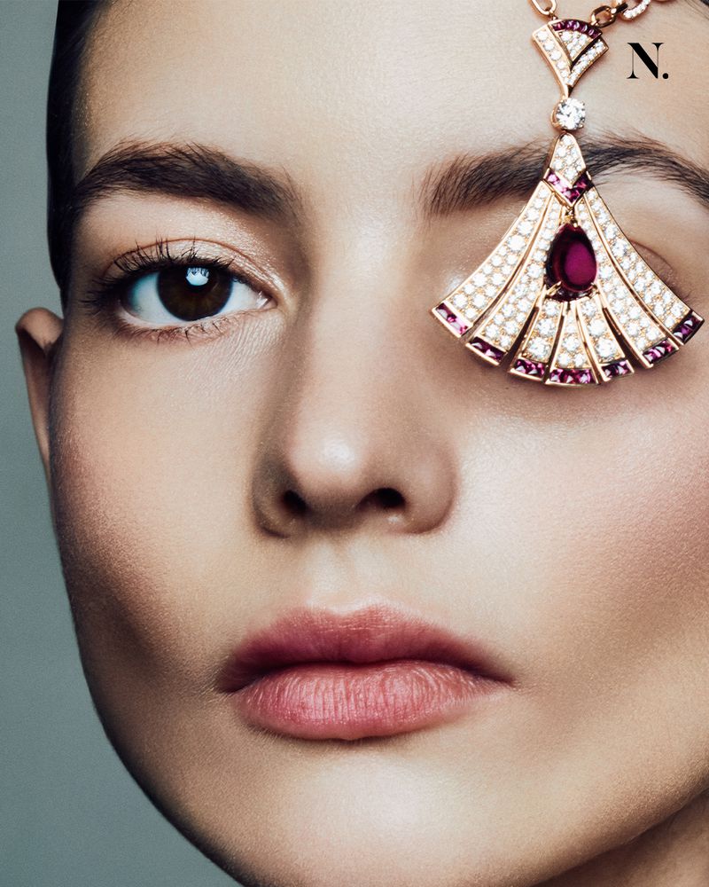 Close-up of a woman's face with a jeweled headpiece. The woman has fair skin, brown hair and eyes, and pink lipstick, gray background.