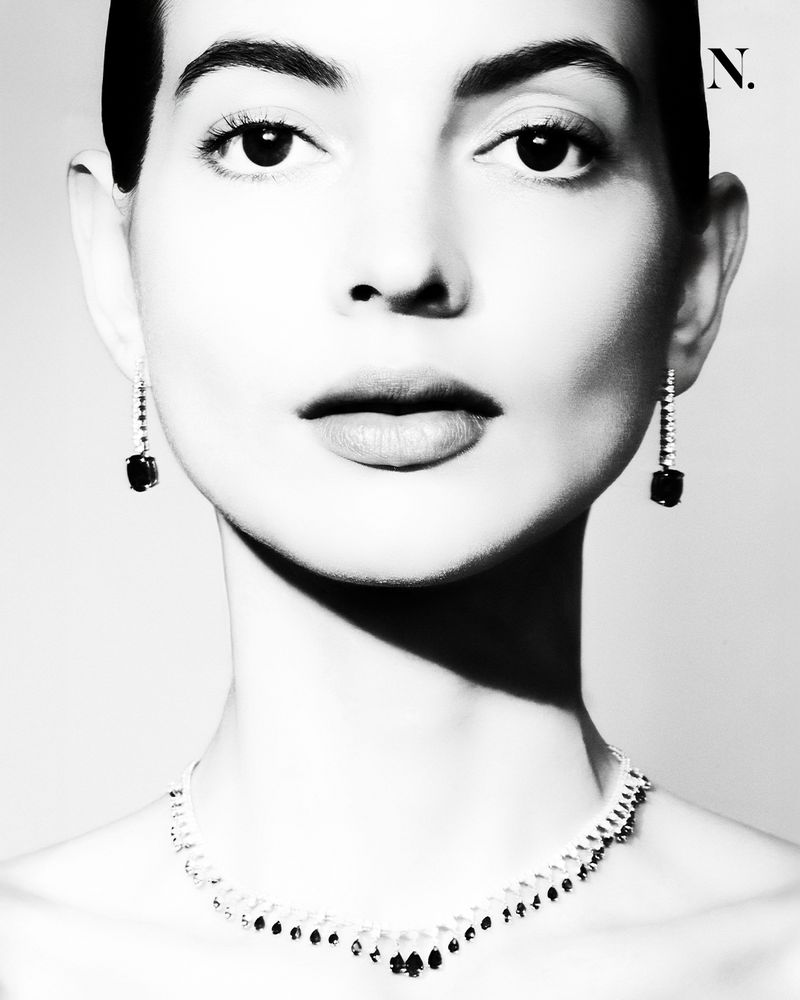Black and white portrait of a woman wearing elegant jewelry including earrings and a necklace, posing in front of a bright background.