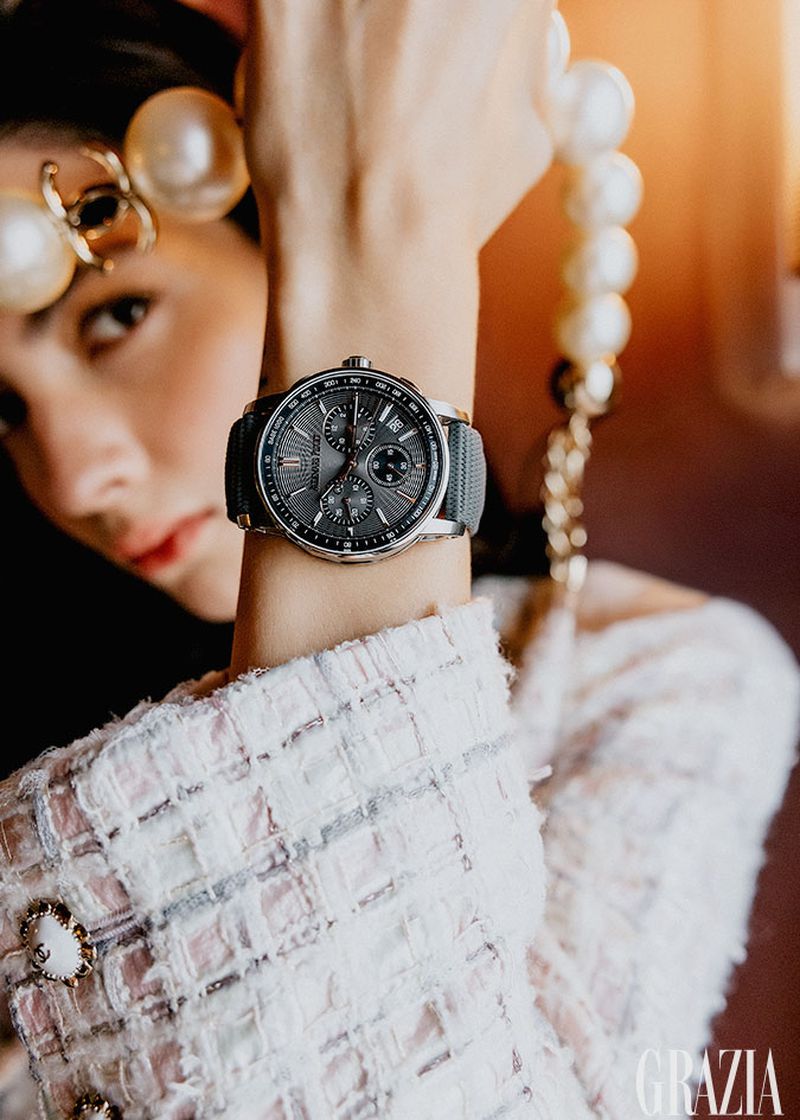 Elegant woman showcases a wristwatch with a gray band, complemented by pearl accessories and a Chanel-style jacket in a high-fashion editorial.