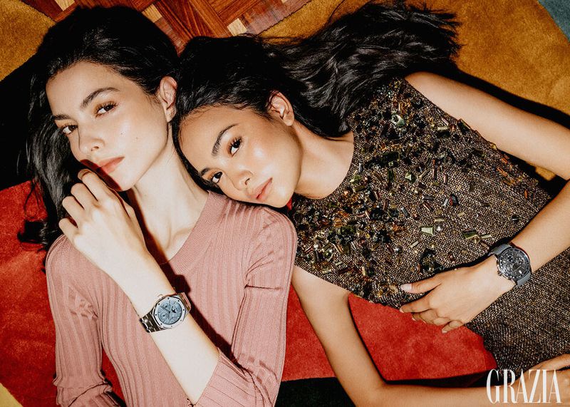 Two women lie on a patterned surface wearing dresses and watches, gazing at the camera in a fashion or beauty editorial setting.