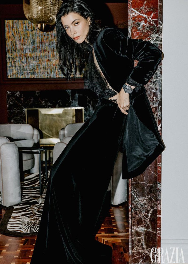 Elegant woman in black velvet dress posing indoors against a marble column and ornate background. Fashion editorial photography.