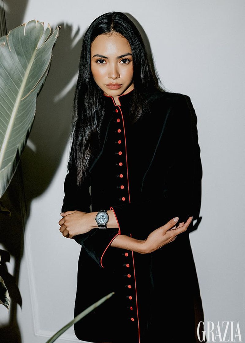 Elegant woman in black jacket with red trim poses in a studio with a plant, showcasing a wristwatch. Grazia magazine logo in the corner.