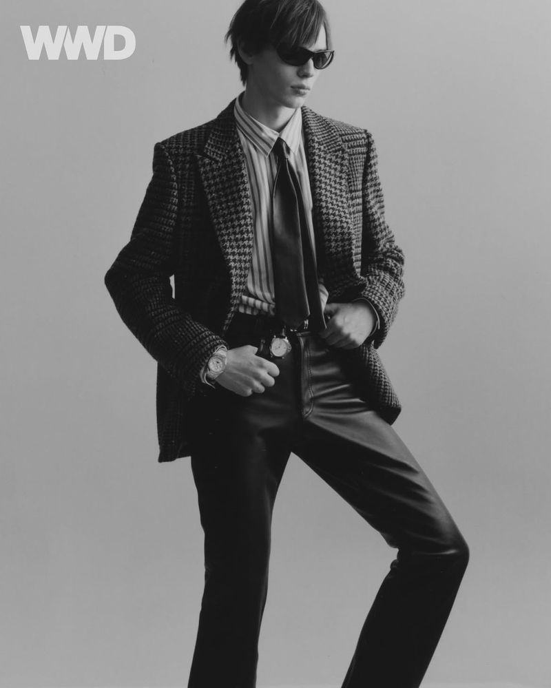 Monochrome fashion portrait of adult male model in blazer, leather pants and sunglasses posing against a grey backdrop.