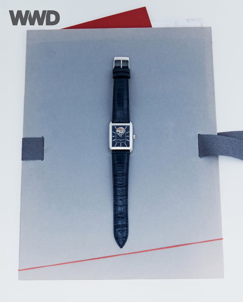 Elegant wrist watch with a square silver case and dark leather band, centered on a gray textured surface with a red accent.