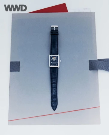 Elegant wrist watch with a square silver case and dark leather band, centered on a gray textured surface with a red accent.