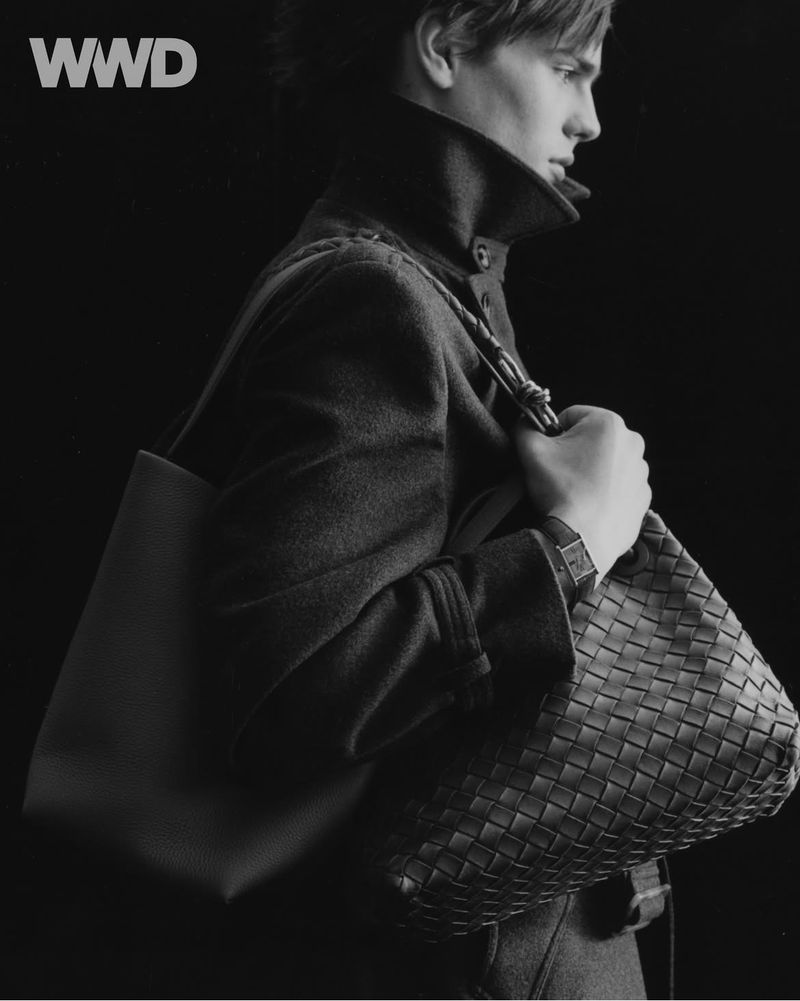 Monochrome fashion portrait of a young man with a dark overcoat and leather tote bag in a studio with black backdrop.