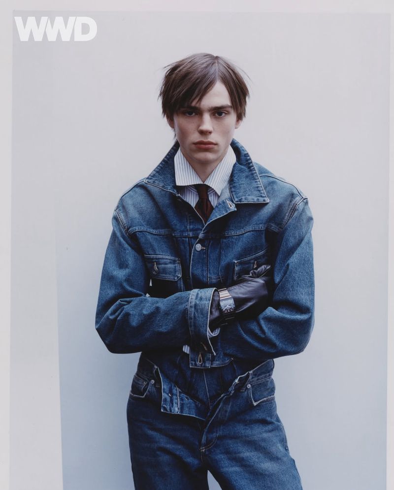 Fashion editorial shot of a young adult man wearing a denim jacket and pants, leather gloves, and tie against a plain background.