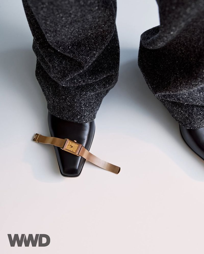 Close-up studio shot of a gold wristwatch on a black loafer with gray tweed pants and a white background in a vertical format.