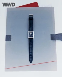 Rectangular silver wristwatch with an embossed black leather strap set against a grey paper presentation sleeve.