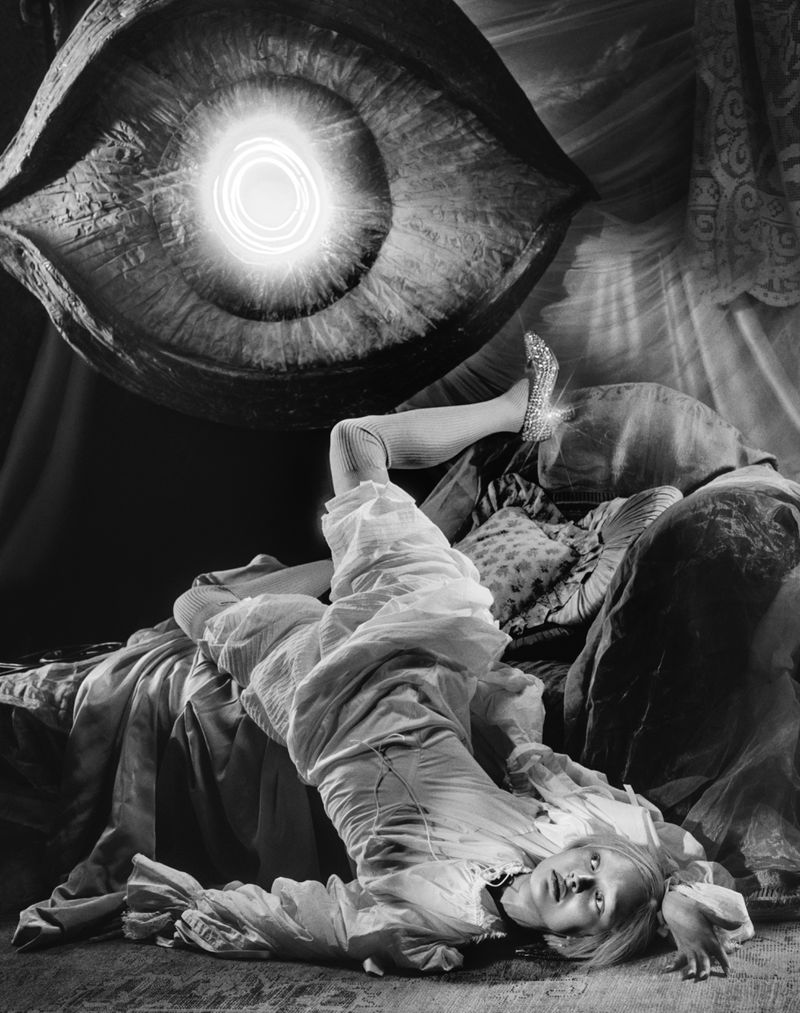 Monochrome studio shot of a woman lying on a bed with an oversized eye and illuminated pupil above her in a surreal, dreamlike scene.