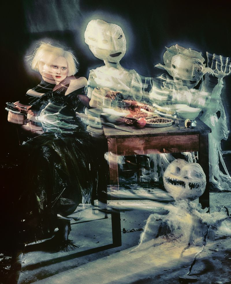 Stylized shot of a fair-skinned woman seated at a table set with food, alongside two ghost-like figures and a smiling pumpkin prop.