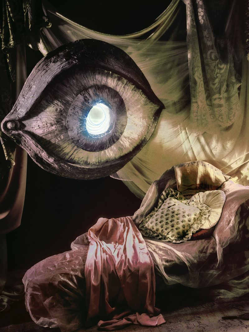Dramatic studio shot of a large eye prop with a bright center, draped with sheer fabrics above a luxurious chaise lounge and decorative pillows.