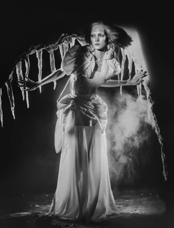 Monochrome studio shot of a model in an elaborate white gown with icicle-like decorations, dramatic lighting, and a smoky background.