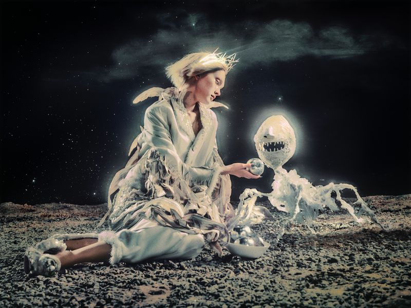 An adult female wearing a crown and a garment with feathers sits facing an odd creature holding an orb in a dark, starry outdoor setting.