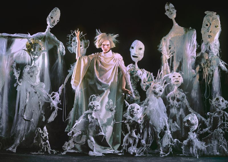 Woman in white robe surrounded by fantastical puppets with stark white costumes and elaborate masks in a theatrical performance.