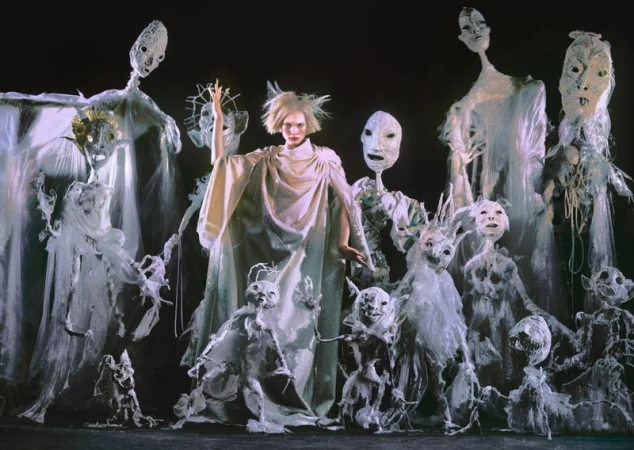 Woman in white robe surrounded by fantastical puppets with stark white costumes and elaborate masks in a theatrical performance.