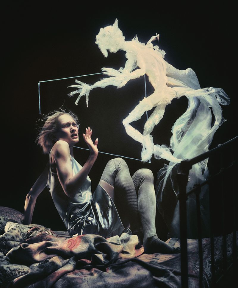 A woman with fair skin on a bed looks scared at a ghost-like figure with outstretched hands hovering over her in a dimly lit space.