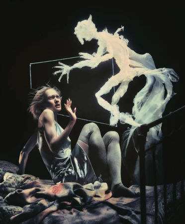 A woman with fair skin on a bed looks scared at a ghost-like figure with outstretched hands hovering over her in a dimly lit space.