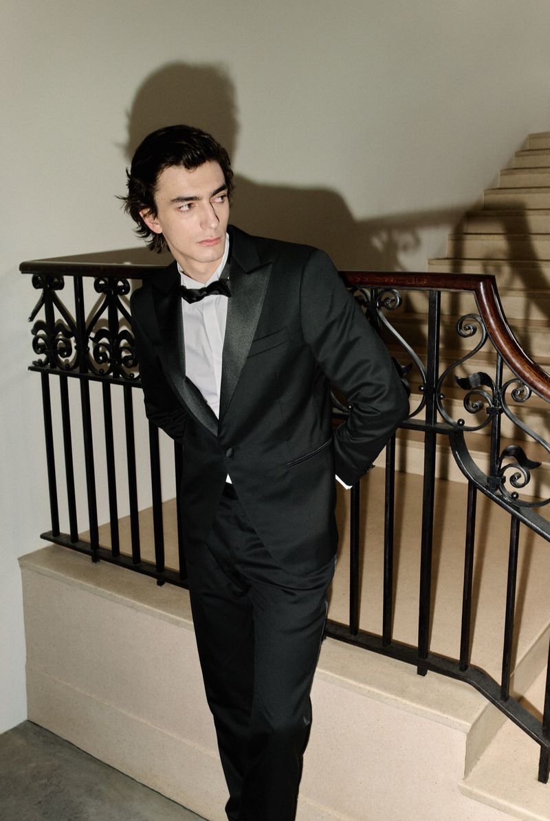 Portrait of a young adult male in a black tuxedo with a bowtie, posing indoors near a staircase with ornate railings. Classic style.