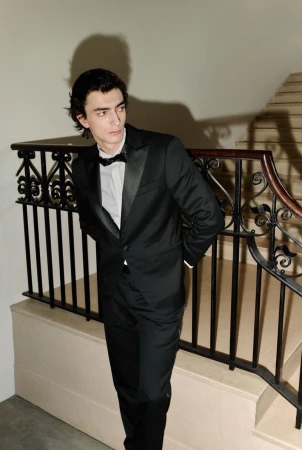 Portrait of a young adult male in a black tuxedo with a bowtie, posing indoors near a staircase with ornate railings. Classic style.