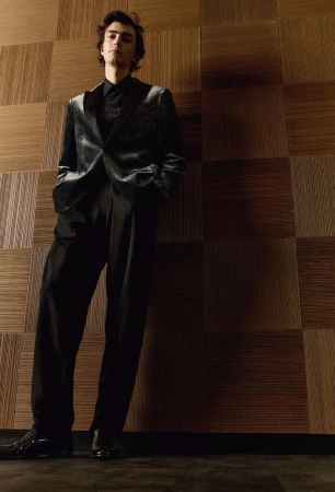 A young adult man in a dark suit and shiny black shoes stands in front of a textured wooden wall looking at the camera.
