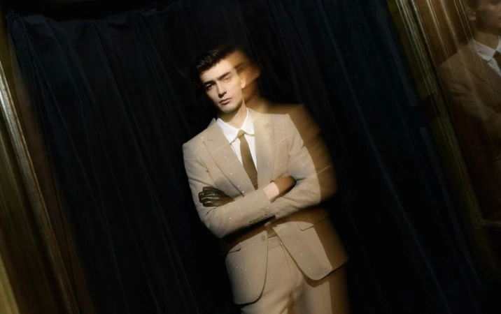 Man in beige suit poses in front of black curtain, arms crossed, lit by spotlights in a close studio shot. Fashion editorial.