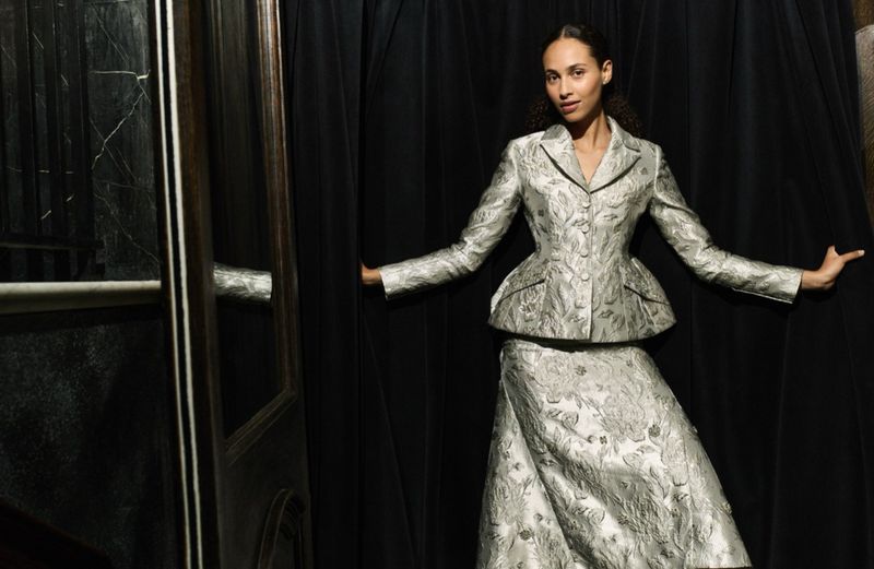 A woman in a silver patterned suit poses, standing between a black curtain and a dark door, in a studio-lit fashion portrait.