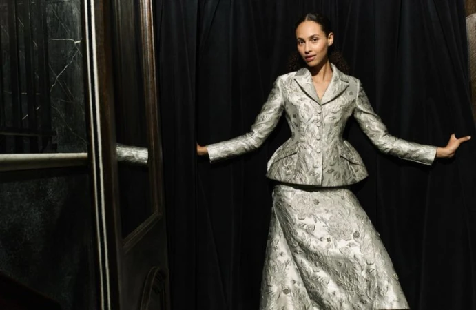 A woman in a silver patterned suit poses, standing between a black curtain and a dark door, in a studio-lit fashion portrait.