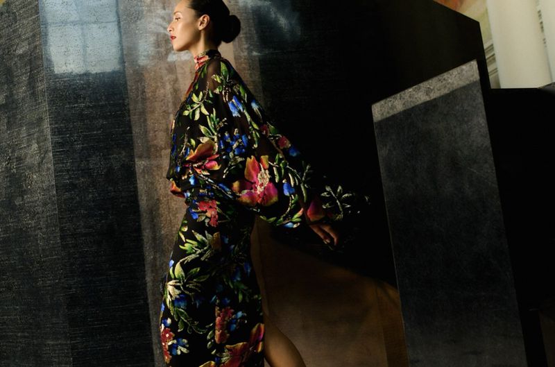 Adult woman in floral print dress and bun hairstyle posing in a studio with gray wall and dark stone blocks.