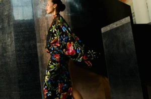 Model Anissa Bakhti in a black floral-patterned dress posing in profile beside tall dark marble panels.