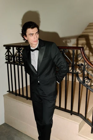 Model Gena Malinin wearing a tailored black tuxedo and bow tie leans against a wrought-iron staircase railing.