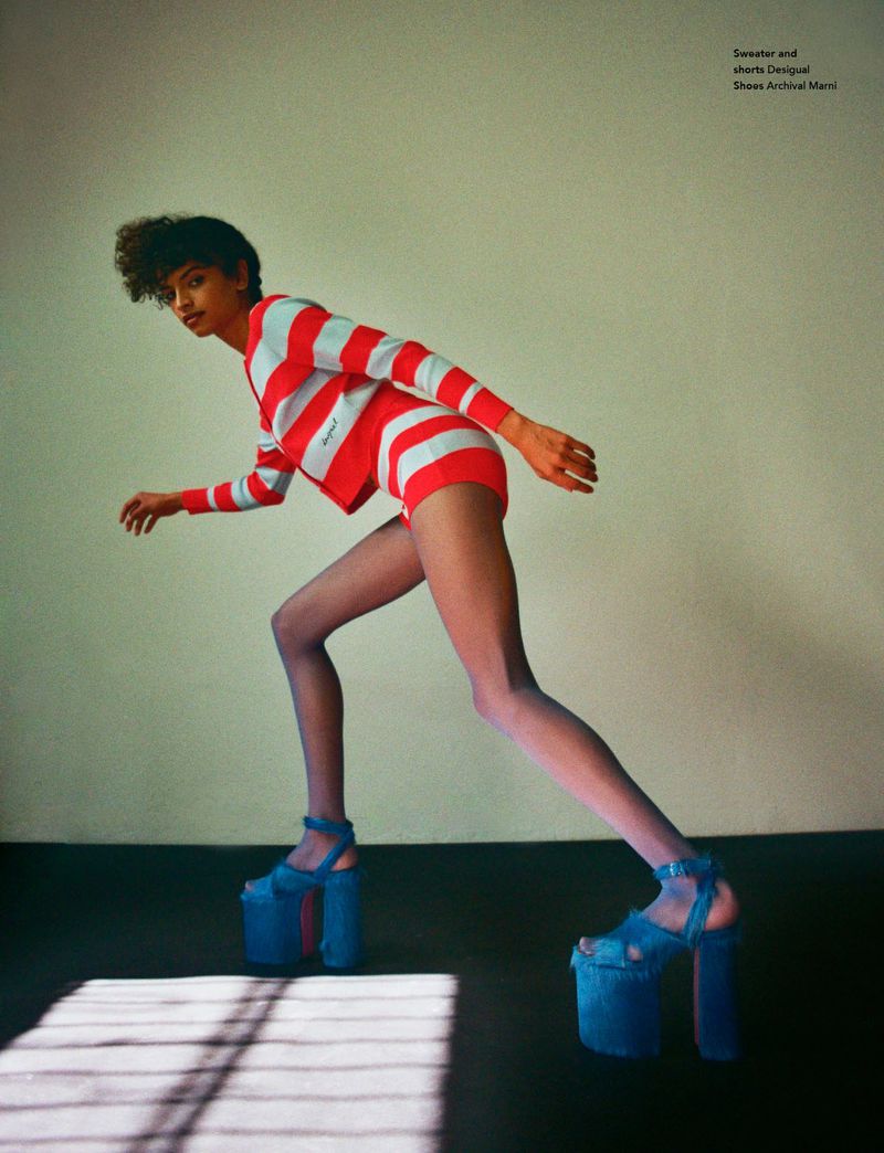 Fashion shot of a woman in red and white striped shorts and top with blue platform heels, posing in a light-walled studio.