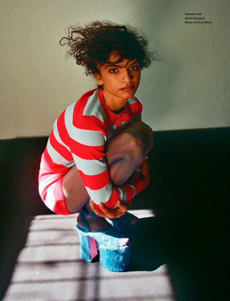 Woman poses crouching indoors wearing a red and white striped shirt, pink shorts, and blue platform shoes with faux fur trim.