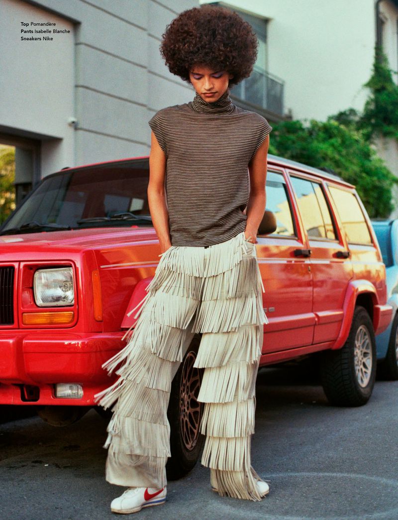 A person with an afro stands next to a red SUV, wearing a brown striped top and fringed beige pants with white sneakers on an asphalt street.